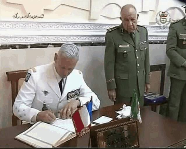 a man in a white uniform is signing a book while another man stands behind him