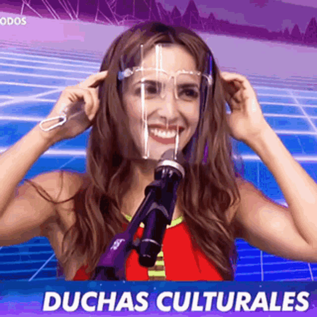 a woman wearing a face shield and glasses stands in front of a microphone with duchas culturales written below her