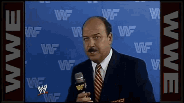 a man in a suit and tie is talking into a microphone in front of a wall that says wwe on it