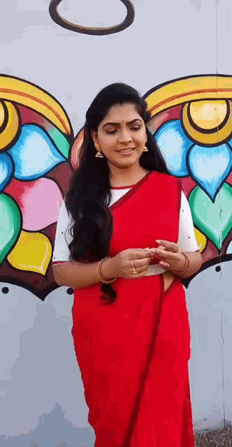 a woman in a red saree is standing in front of a colorful painting on a wall .