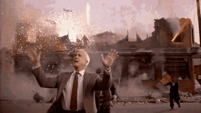 a man in a suit and tie is standing in front of a destroyed building with fireworks exploding in the background
