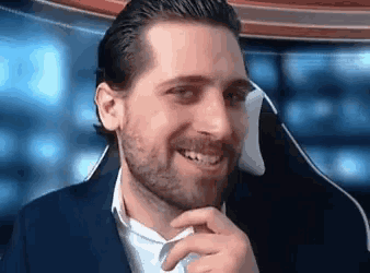 a man with a beard is smiling while sitting in a chair with his hand on his chin .
