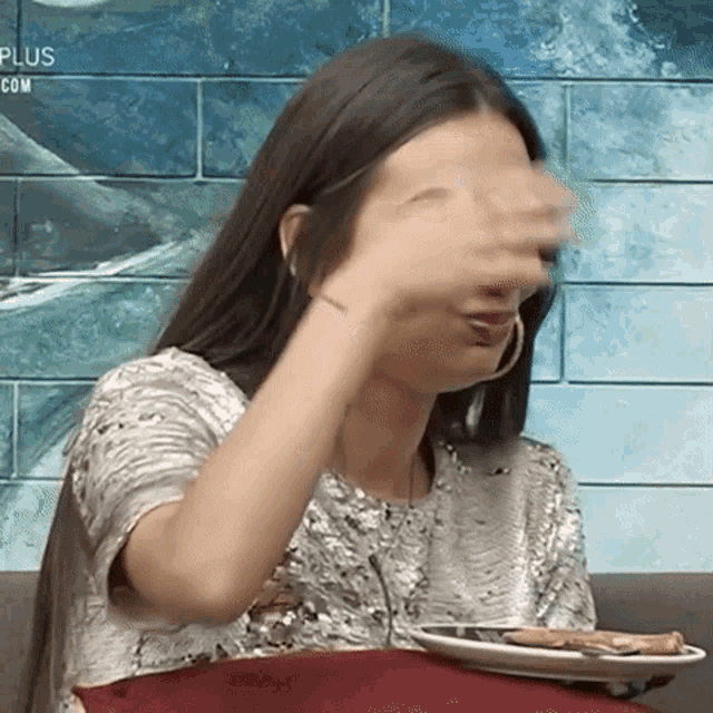 a woman covering her face with her hand while holding a plate of food in front of a sign that says plus.com