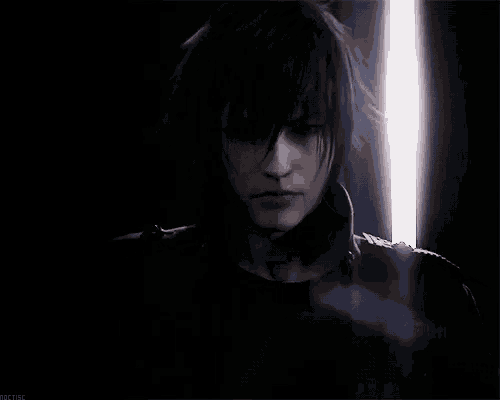 a man with long black hair is standing in a dark room with a light behind him