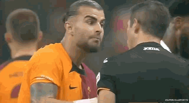 a man in an orange nike shirt is talking to another man in a black joma jersey .
