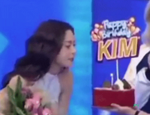 a woman is blowing out a candle on a birthday cake in front of a sign that says `` happy birthday kim '' .