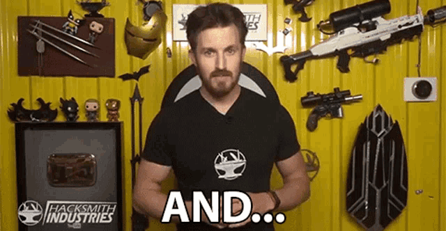 a man in a hacksmith industries shirt is standing in front of a yellow wall holding a gun .