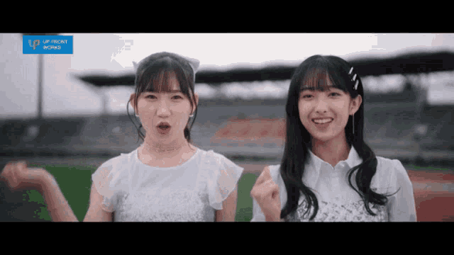 two girls are standing next to each other in front of an empty stadium and a blue sign that says up front world