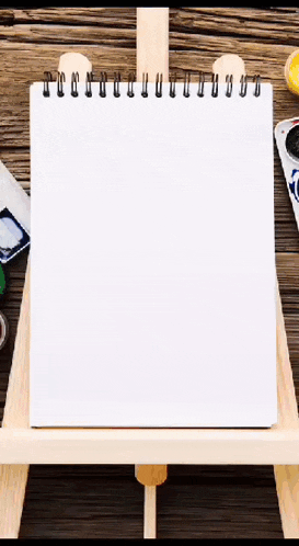 a notebook is sitting on a wooden easel next to a palette of paints