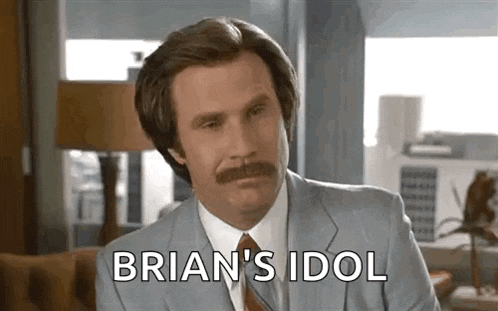 a man with a mustache and a suit is sitting on a couch and saying brian 's idol .