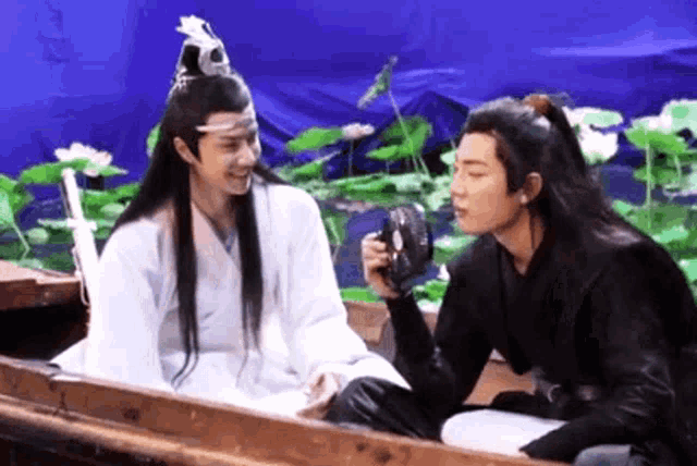 two men are sitting in a boat talking to each other and one is holding a camera .