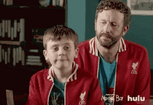 a man and a boy are sitting next to each other . the boy is wearing a liverpool jacket .
