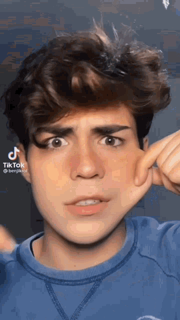 a young man in a blue shirt is making a funny face while holding his hand to his face .