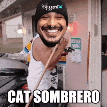 a man wearing a black beanie and a white tank top is smiling and holding a broom and says cat sombrero