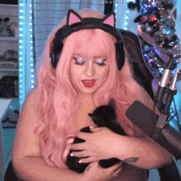 a woman with pink hair and headphones holds a cat