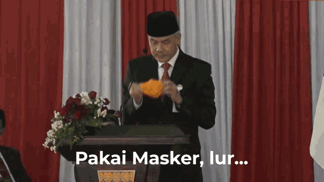 a man in a suit and tie stands at a podium holding a mask and says pakai masker