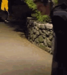 a man in a black shirt stands on a sidewalk next to a stone wall