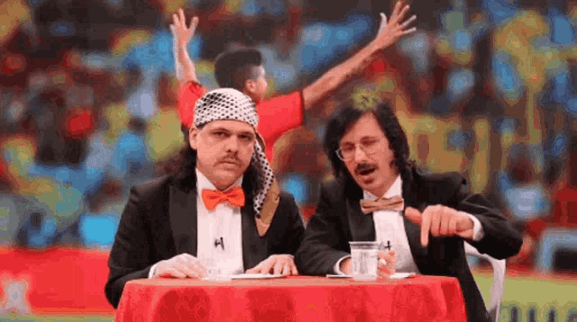 two men in tuxedos sit at a table with the letter h on the table