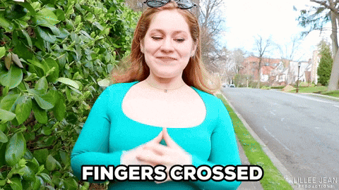 a woman in a blue shirt says fingers crossed in front of a bush