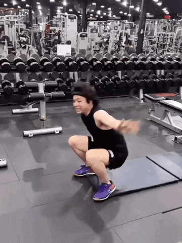 a man is squatting on a mat in a gym with a lot of dumbbells behind him .