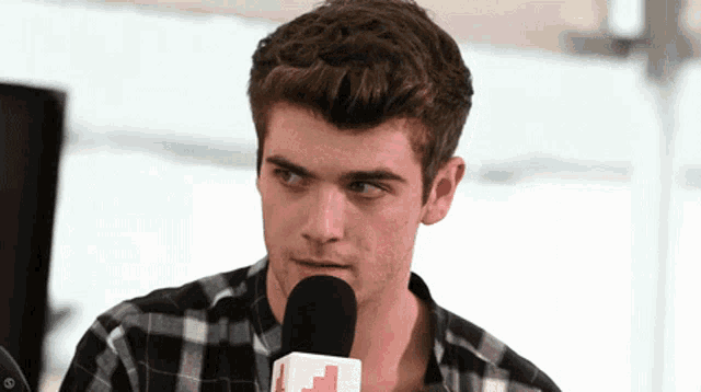 a man in a plaid shirt talks into a microphone with a red cross on it