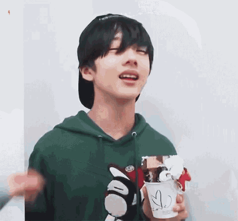 a young man in a green hoodie is holding a cup of ice cream and smiling .