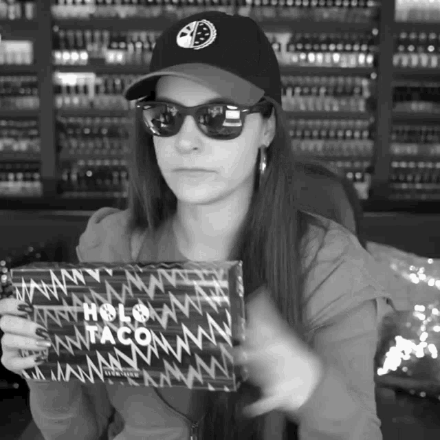 a woman wearing sunglasses and a baseball cap holds a box that says holo taco