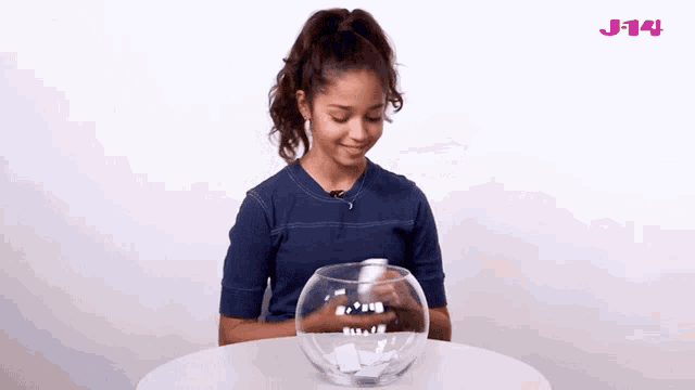a girl is sitting at a table with a bowl of paper in front of her that says j-14 on the top