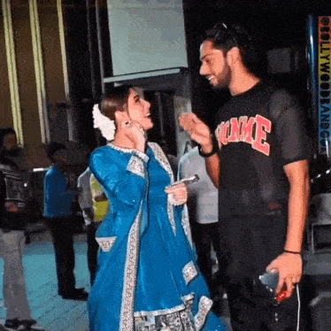 a woman in a blue dress is talking to a man wearing a t-shirt that says ' hollywood ' on it