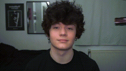 a young man with curly hair looks at the camera in front of a mirror
