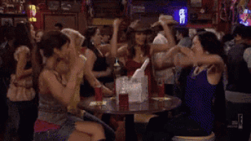 a group of women are sitting at a table in a bar raising their arms in the air .