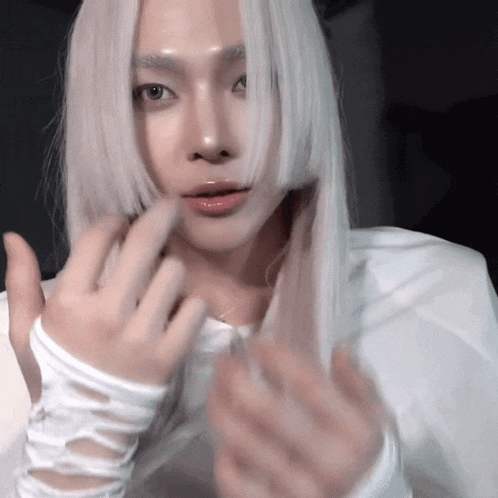a woman with long white hair is wearing white gloves and making a heart shape with her hands .