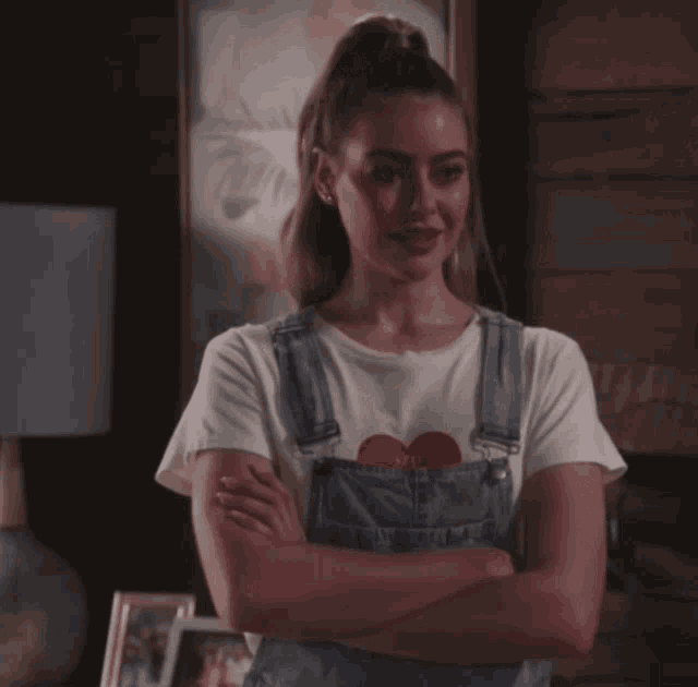 a woman with her arms crossed wearing overalls and a white shirt with a red heart on it