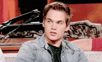 a young man in a denim jacket is sitting on a couch with a crossbow in the background