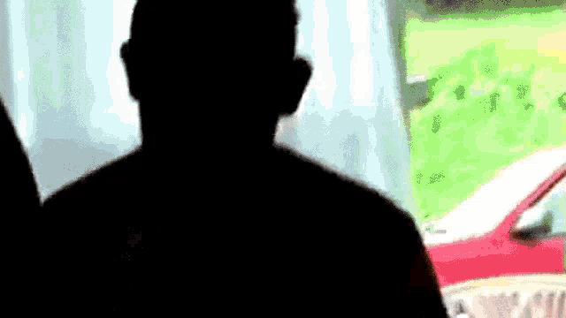 a silhouette of a man in front of a window with a red car in the background .