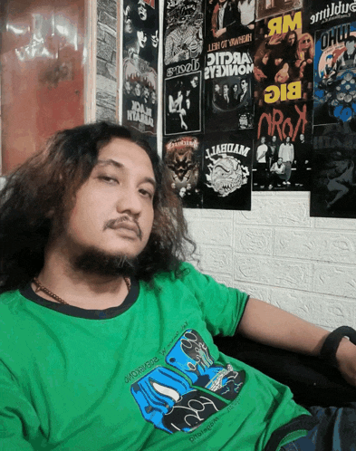 a man in a green shirt is sitting in front of a wall with posters on it including one that says big oryx