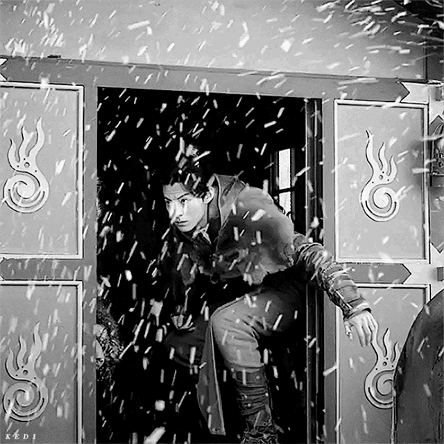 a black and white photo of a man standing in a doorway with snow falling and the letters k & d on the bottom right