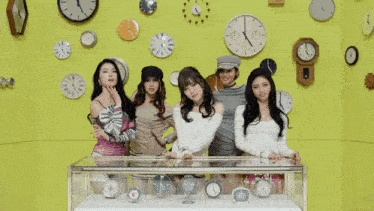 three women are dancing in front of a yellow wall with clocks on it and one of the clocks shows the time as 10:10