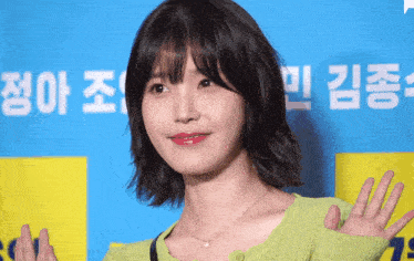 a woman in a green sweater is waving in front of a blue and yellow sign that says ' a ' on it