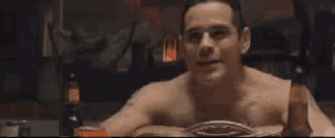 a shirtless man is sitting in a dark room and looking at the camera .