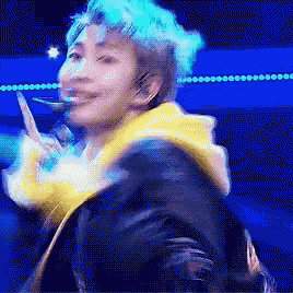 a man with blue hair is wearing a yellow jacket and giving a peace sign .