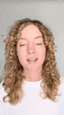 a woman with curly blonde hair is wearing a white shirt and smiling with her eyes closed .