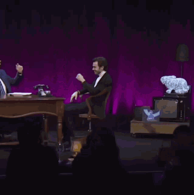 a man in a suit sits on a stool talking to another man