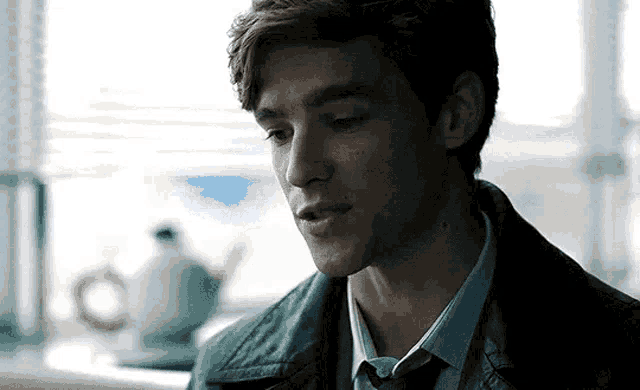 a young man in a leather jacket and tie is sitting at a table in front of a window .
