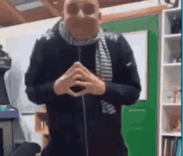 a man wearing a scarf and sunglasses is standing in a room with his hands folded .