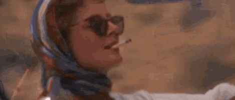 a woman wearing sunglasses , a scarf and a ring is waving her hand .