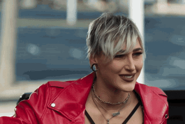 a woman with short blonde hair is wearing a red leather jacket and necklaces