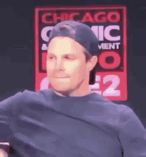 a man wearing a baseball cap and a sweater is standing in front of a sign that says chicago comic con .