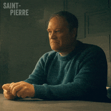 a man in a blue sweater sits at a table with saint-pierre written on the wall