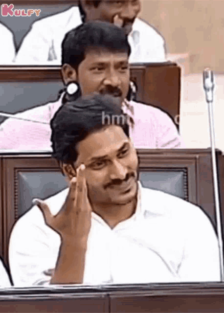 a man with a mustache is sitting in a row of chairs with his hand on his forehead .